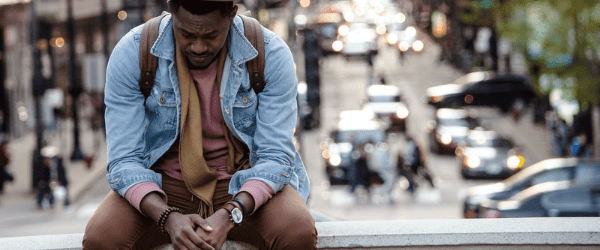 depressed black man sits on a the ledge of a bridge over looking a busy street